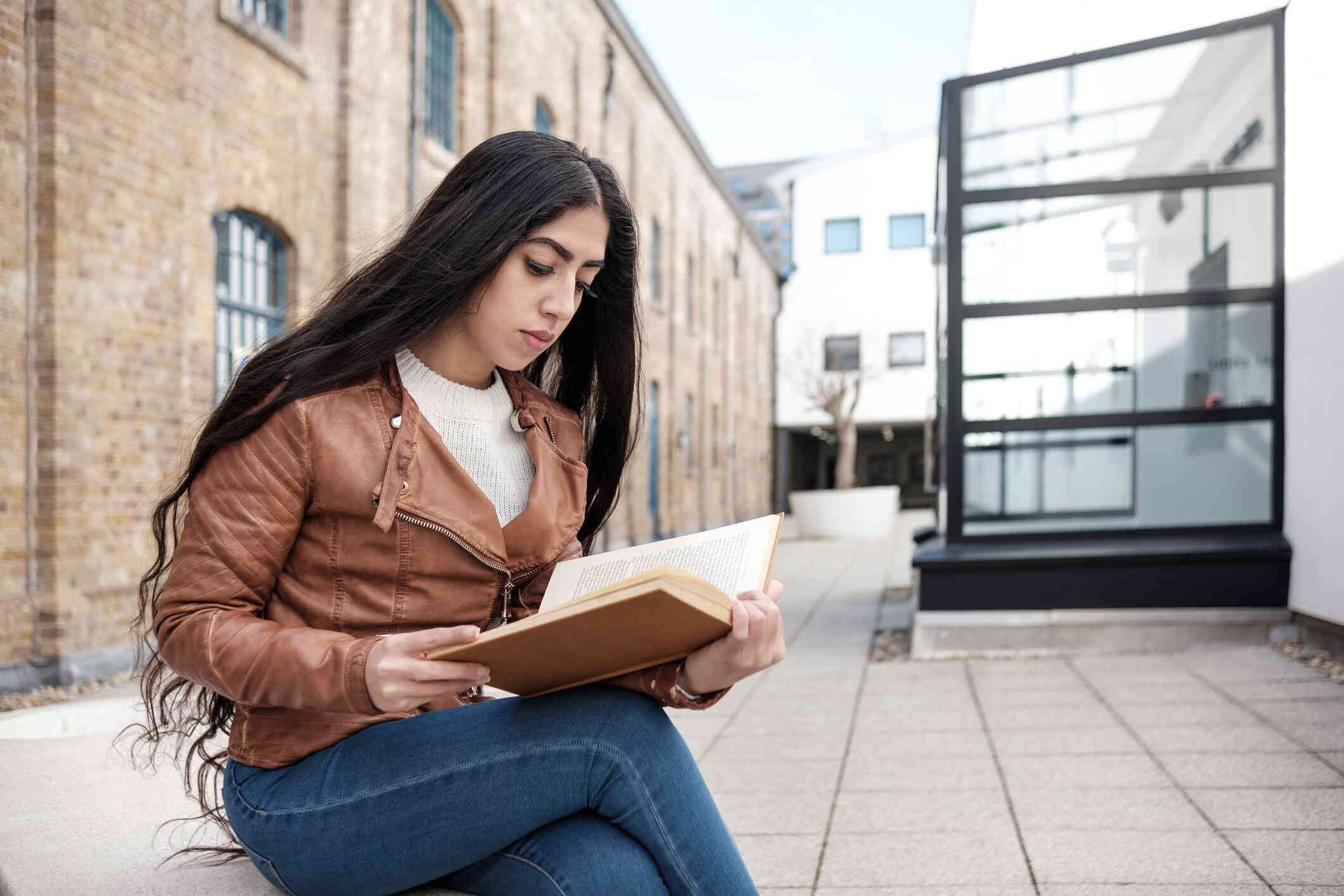 A woman in a brown leather jacket sits outside on a bench and reads from a book.